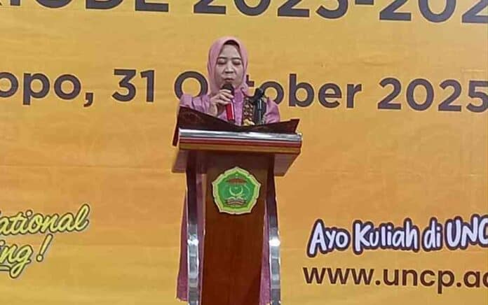 Karyawan wanita berbaju batik dan hijab sedang berbicara di depan mikrofon di acara resmi universitas.