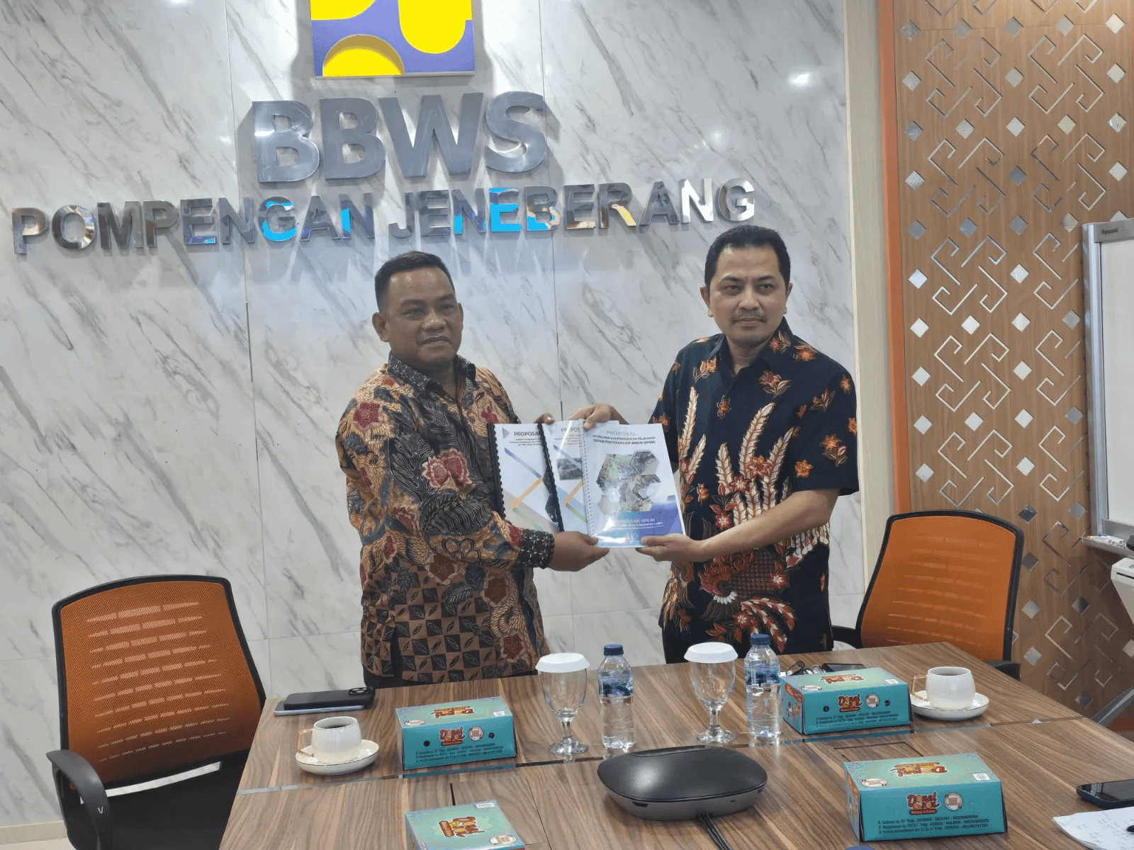 Pertukaran dokumen antara dua pria di ruang rapat, dengan latar belakang logo BWS BBWS Pompengan Neberang.